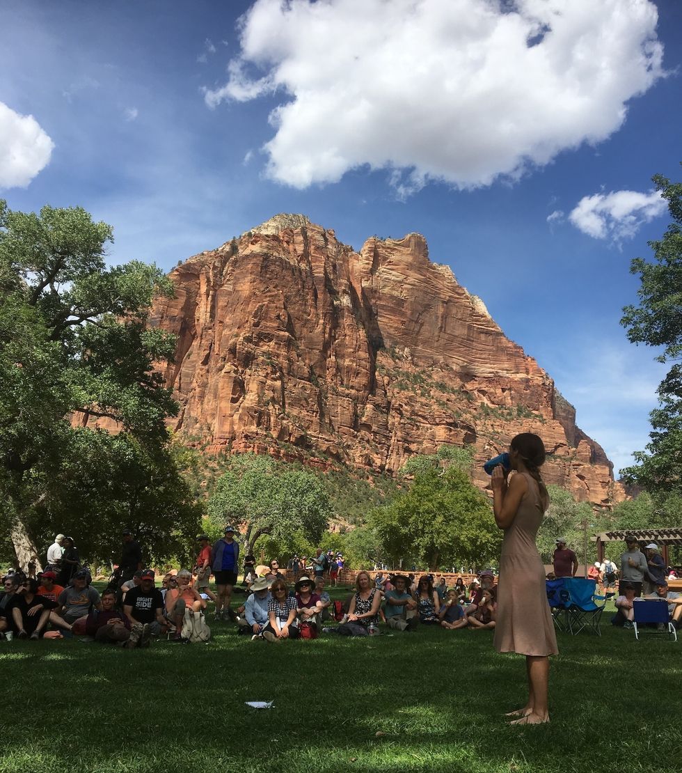 Longoria sips from a water bottle will standing on a grassy lawn. A large group of park visitors are seated in the grass watching.