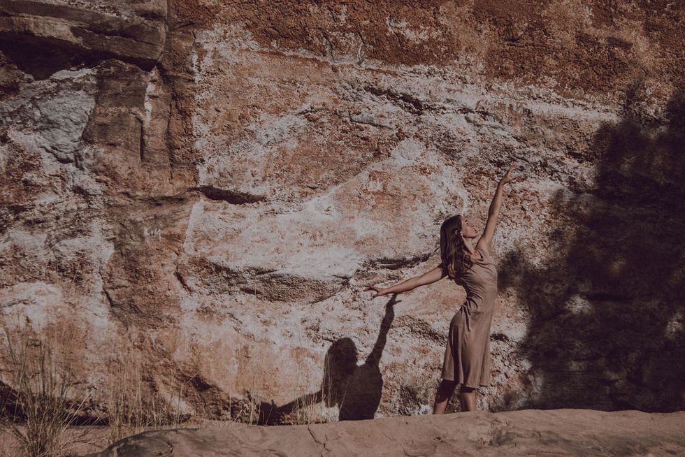 Longoria, with long brown hair and a tan dress, blends in with her rocky background, dancing with arms raised.