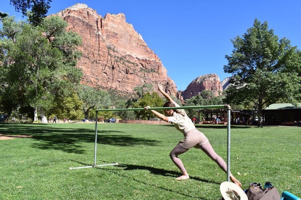 Longoria in a cambru00e9 forward as she pliu00e9s on her supporting leg and her working leg extends in a tendu derriere. She is at the ballet barre on the lawn in front of a vast mountain view.