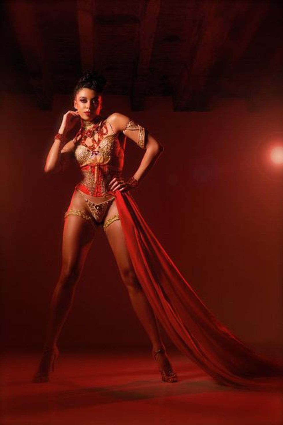 A woman staring intensely at the camera, with moody red lighting. She is wearing a decorative bikini style outfit, with a draped cloth running from her hip.