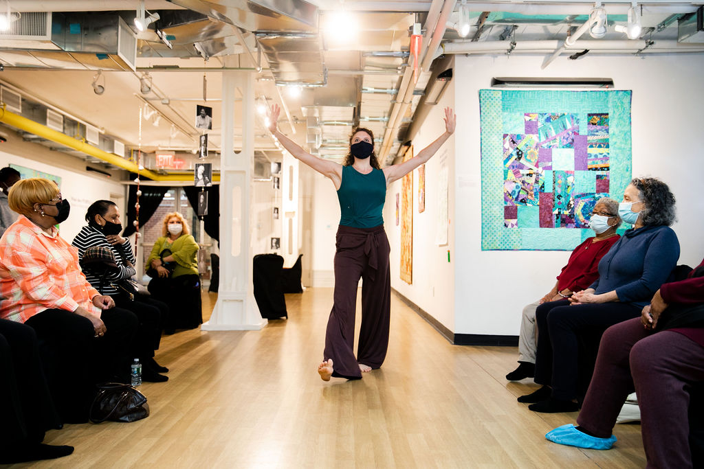 A barefoot dancer strides forward with her arms outstretched beside her ears, palms pressing out. A small handful of older audience members sit, watching raptly, on either side of the space. Everyone wears face masks covering their noses and mouths.
