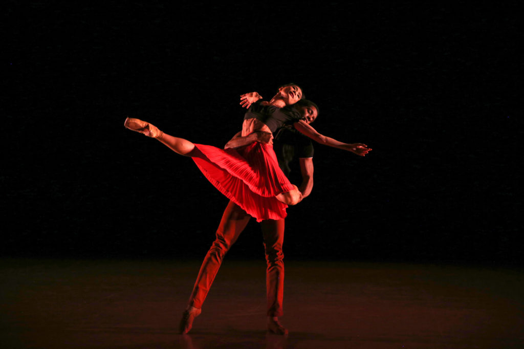 A male dancer lifts a female dancer in a red dress, holding her at the waist and on one knee. Her other leg extends in front of her and she reclines into him her arms to the side and back.
