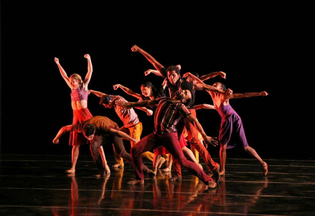 A group of about 10 dancers cluster onstage in different standing positions, arms raised and fists clasped.