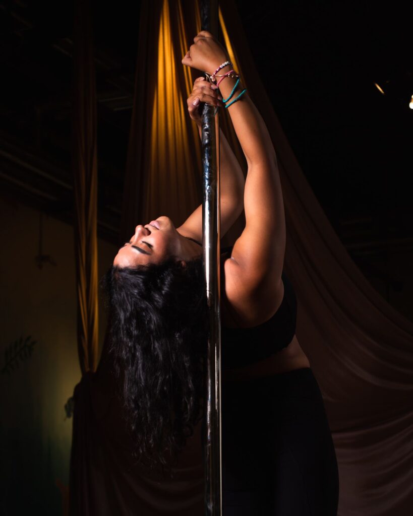 Sharoubim, a woman with long dark hair wearing a black top and pants, leans backward while holding onto a pole, her face and arms illuminated by a spotlight.